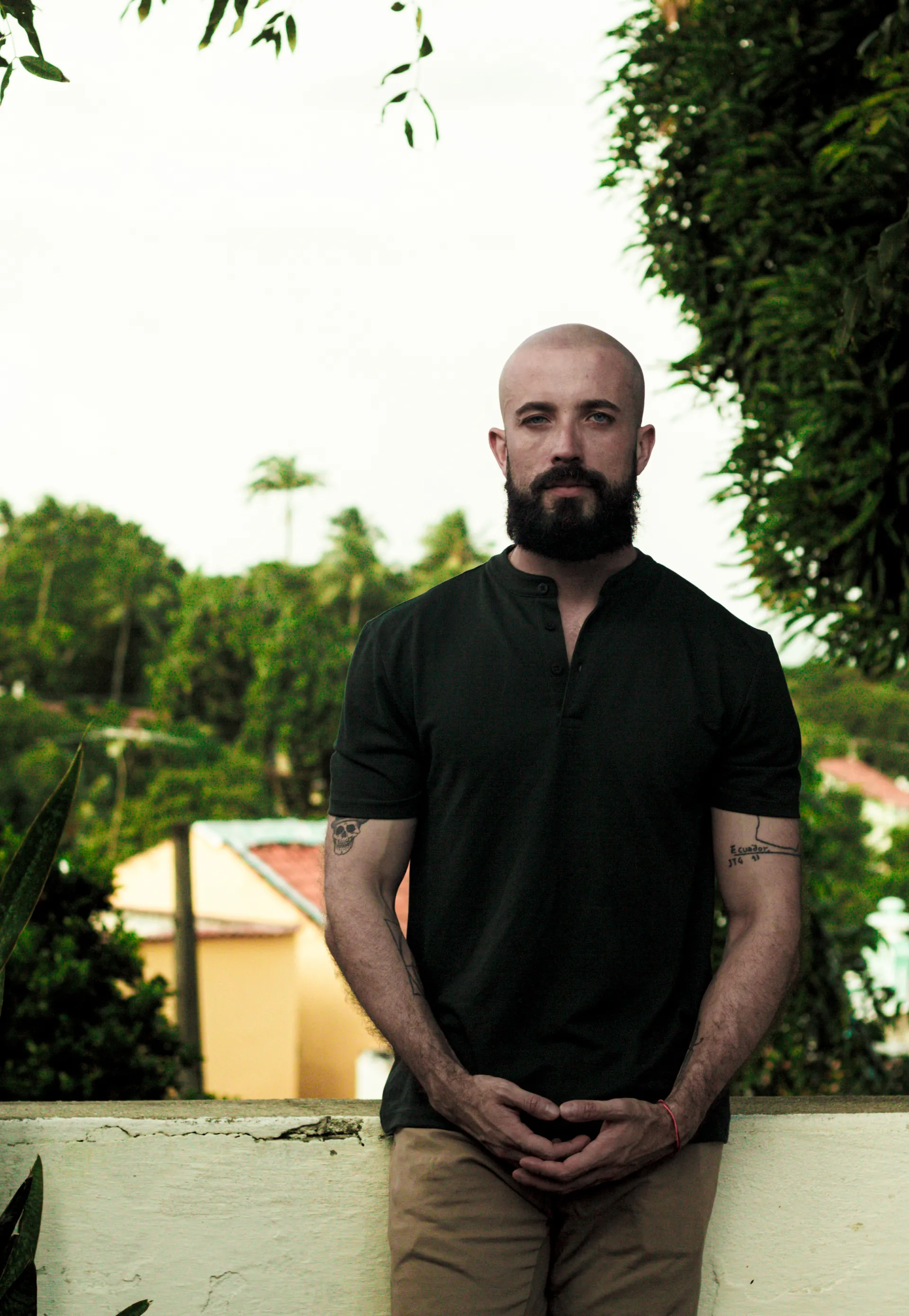 Homem careca e com barba usando camiseta preta, em pé ao ar livre com árvores e casas ao fundo.